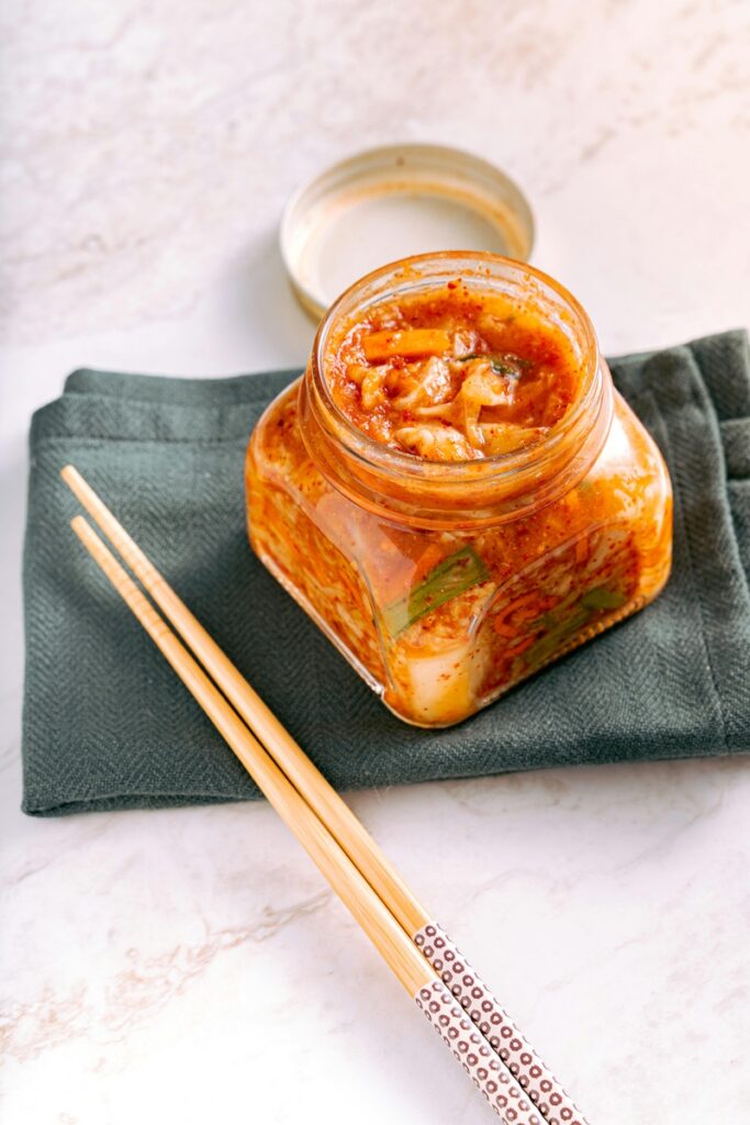 “Glass jar filled with traditional Korean kimchi placed on a cloth with chopsticks beside it.”