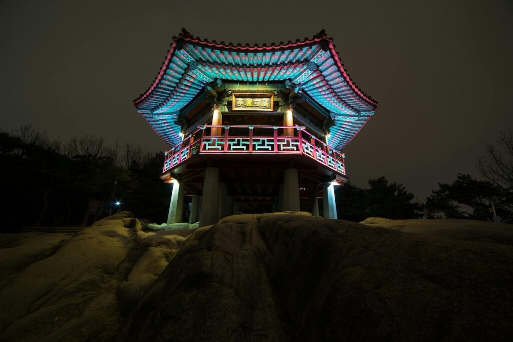 A night view of a traditional pavilion on Achasan Mountain in Seoul, softly illuminated under the stars with the city skyline glowing in the distance.