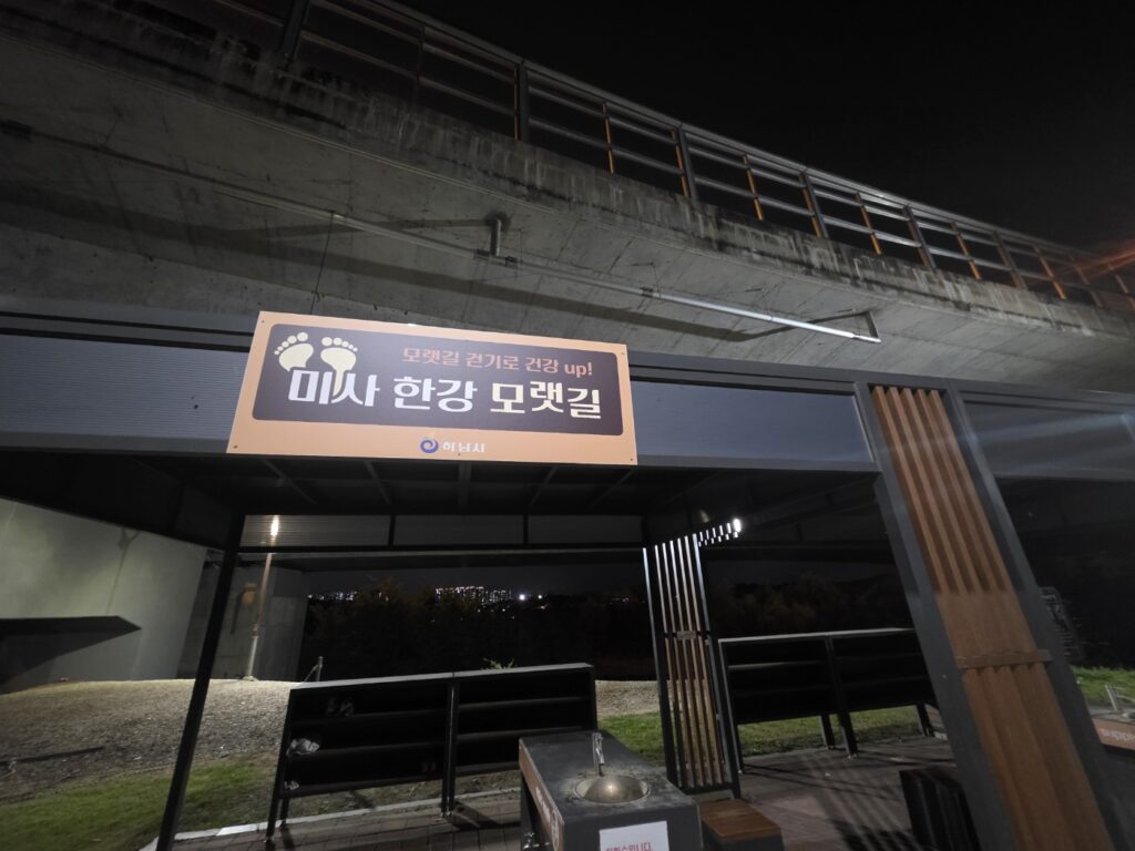 A person walking barefoot on a quiet sand path along the Han River at night, feeling the cool texture of the riverbank beneath soft lights.