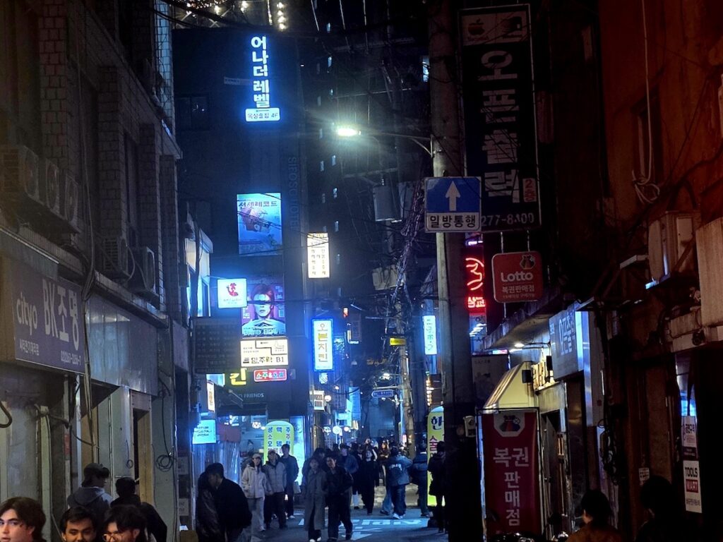 best-seoul-night-road-euljiro-blue-neon-alley A high-contrast night shot of an Euljiro alleyway along the Best Seoul Night Road. Blue neon signs for "어나더레벨" (Another Level) and "오븐치킨" (Oven Chicken) cut through the darkness, while people walk through the narrow space under a tangled mess of overhead power lines and "Lotto" signs.