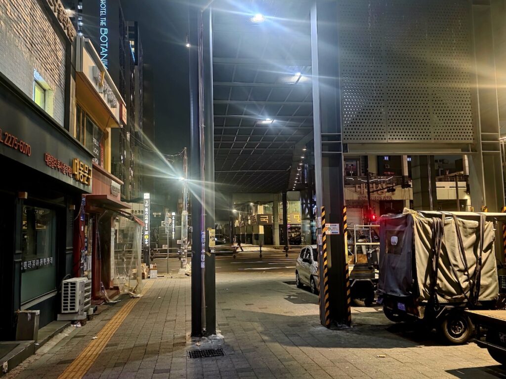 best-seoul-night-road-euljiro-daelim-plaza A wide-angle shot of the modern structural elements of Daelim Plaza (Sewoon Plaza) in Euljiro. The cold, metallic beams and gridded ceiling contrast with the warm, aging signs of "대동문구" (Daedong Stationery) and "국도조명" (Kukdo Lighting) along the Best Seoul Night Road.