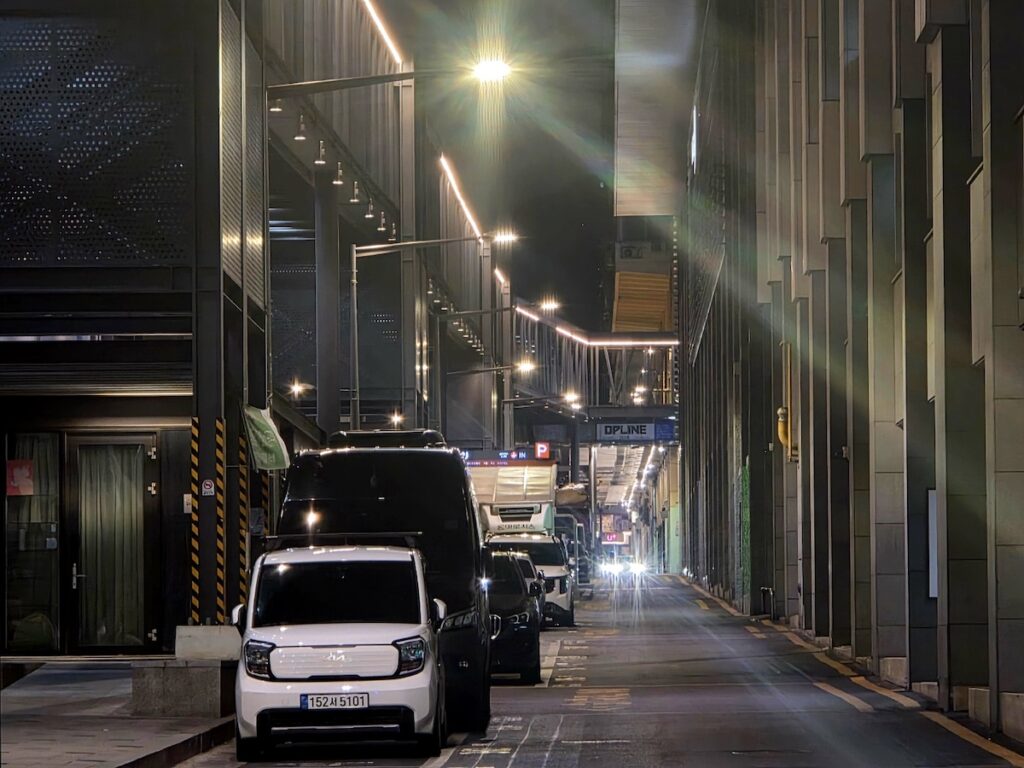best-seoul-night-road-euljiro-modern-corridor A wide-angle night view of a modern industrial corridor in Euljiro. Intense starburst light flares from overhead lamps illuminate a white Kia Ray and several black vans parked along the narrow street, capturing the sleek yet industrial functionalism of the Best Seoul Night Road.