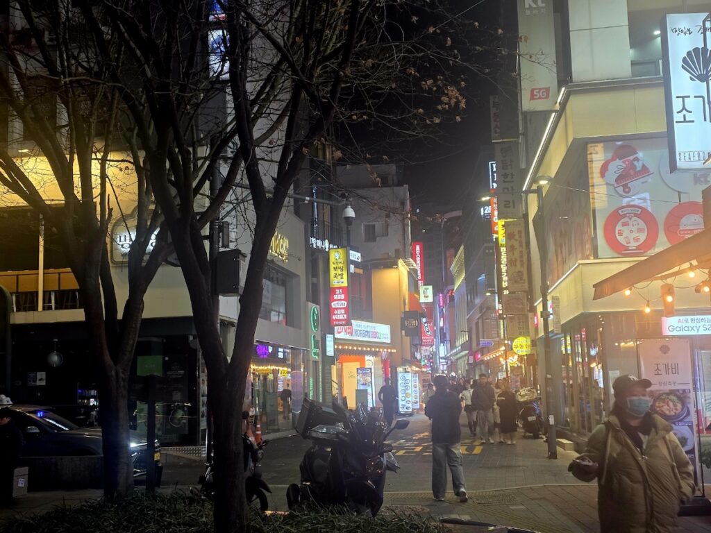 best-seoul-night-road-myeongdong-golden-alley A warm, golden-toned night view of a narrow Myeongdong side street. Bare trees frame the shot, with illuminated signs for "조가비" (Jogabi Seafood) and "Galaxy S24" visible. A person in a heavy winter jacket and mask walks toward the camera, capturing the chilly yet vibrant atmosphere of the Best Seoul Night Road.
