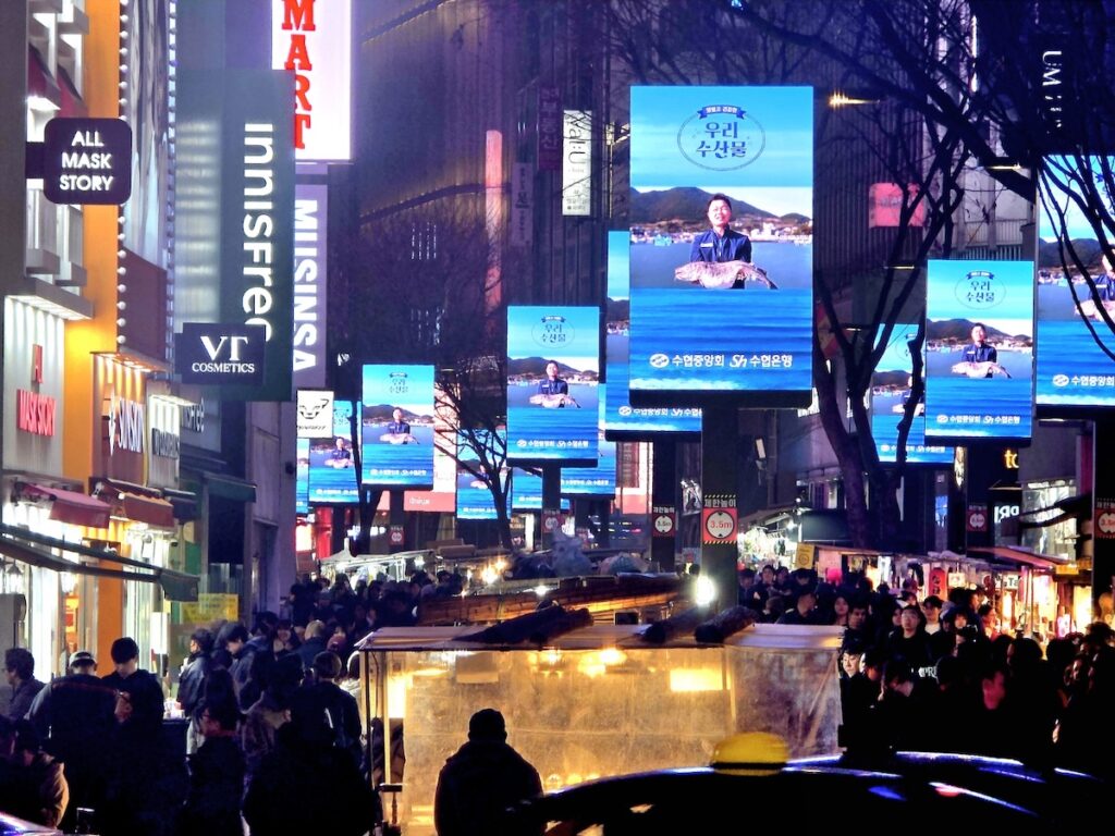 A high-resolution photo taken at night from inside the Lotte Hotel, looking down at the Best Seoul Night Road starting point in Myeongdong. The view is dominated by intense, colorful LED billboards displaying generic Korean text and high-rise office buildings, captured with the unique window reflection of the hotel room. It illustrates the futuristic glitz of the modern city before transitioning into historical alleys.