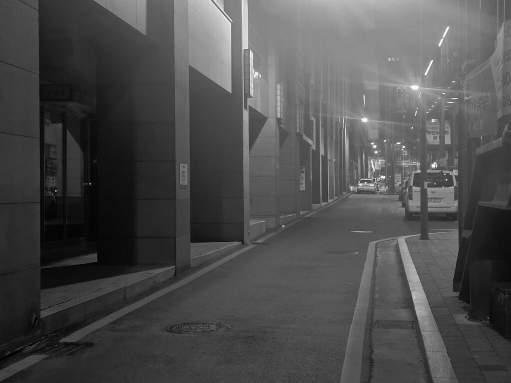 best-seoul-night-road-seoul-monochrome-alley A high-contrast, black-and-white style night shot (or heavy fog effect) of a quiet office alley. The dramatic lighting creates long shadows along the concrete pillars and empty sidewalk, showing the sudden silence of the Best Seoul Night Road away from the neon crowds.