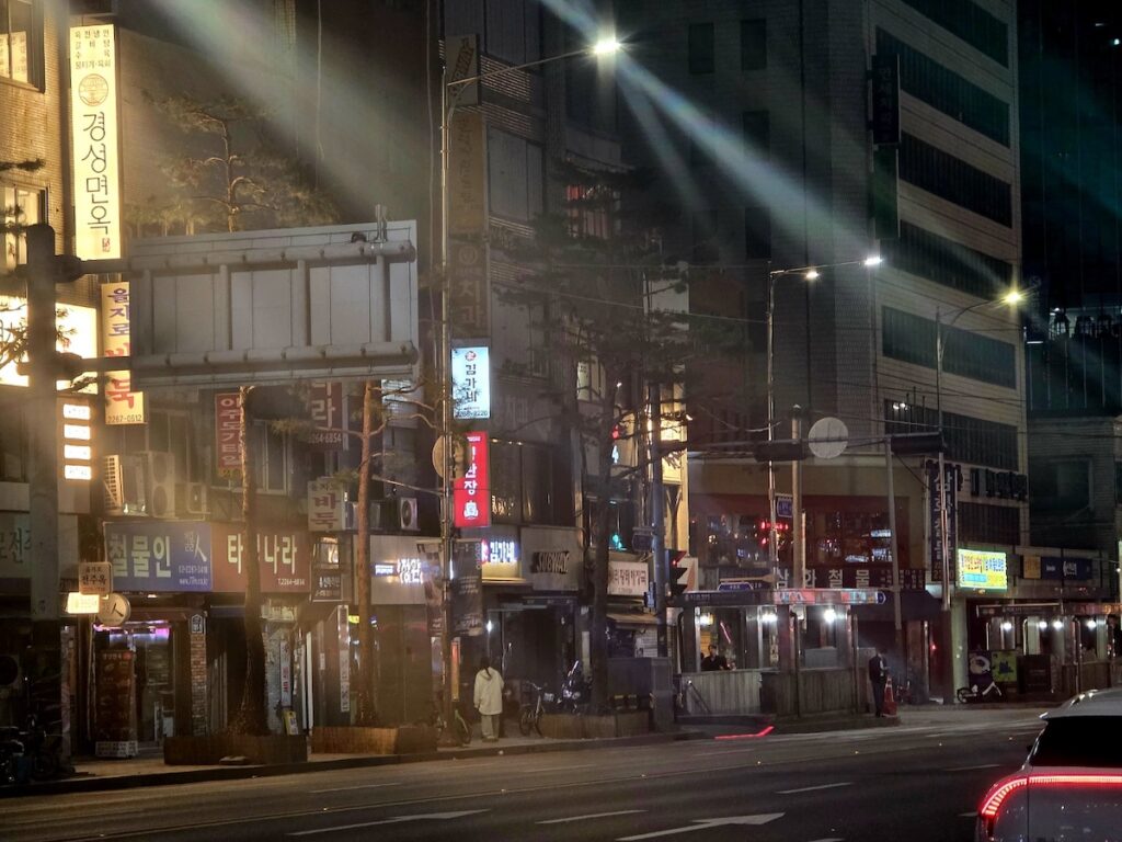 best-seoul-night-road-toegyero-light-streaks A cinematic night shot of Toegye-ro featuring intense light flares from street lamps. The scene includes vintage signs for "경성면옥" (Kyungsung Myeonok) and "김가네" (Kimgane), with the gritty texture of old buildings clashing with the modern light of a passing car's red taillights on the Best Seoul Night Road.