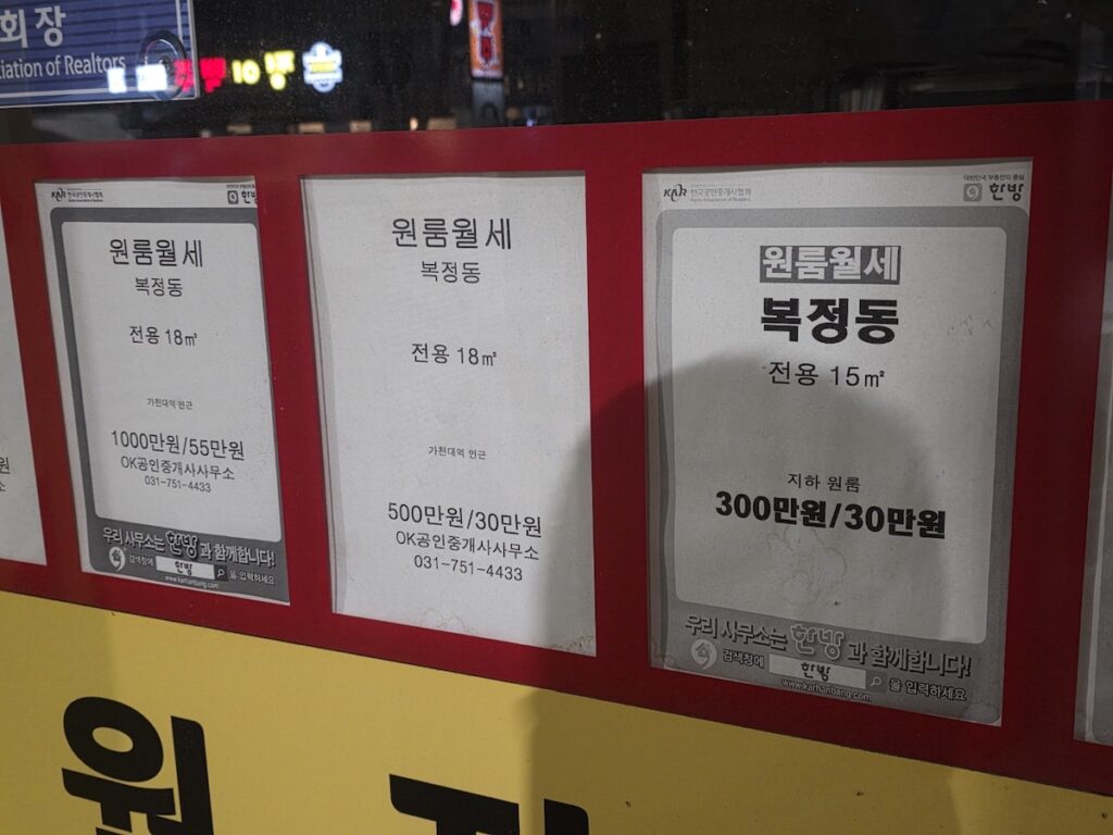 bokjeong-dong-villa-rent-real-estate-listings Close-up of handwritten and printed studio rental listings on a local real estate office window in Bokjeong-dong, showing monthly rates between 300,000 KRW to 550,000 KRW as concrete evidence of the 50% discount compared to Gangnam.