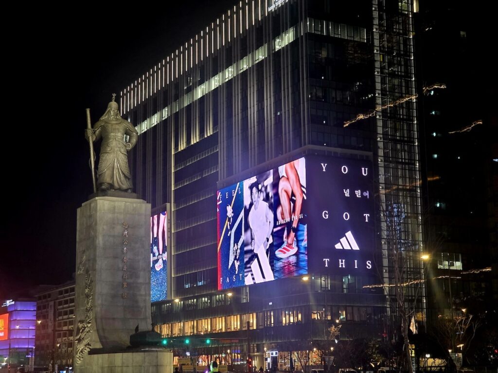 bts-gwanghwamun-locations-2026-adidas-billboard BTS Gwanghwamun Locations 2026 showing the iconic Admiral Yi Sun-sin statue in front of a giant digital billboard featuring an inspirational "YOU GOT THIS" message in Seoul.