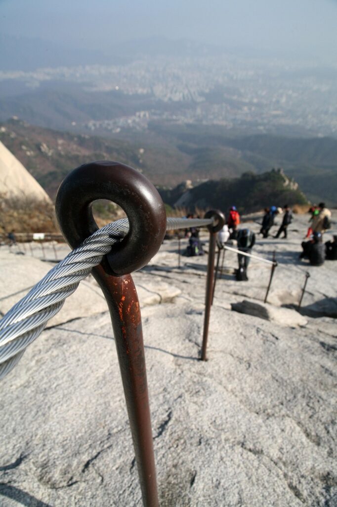 A hiker climbing a rocky trail using a safety rope on Bukhansan Mountain in Seoul, South Korea, surrounded by granite cliffs and clear blue skies.