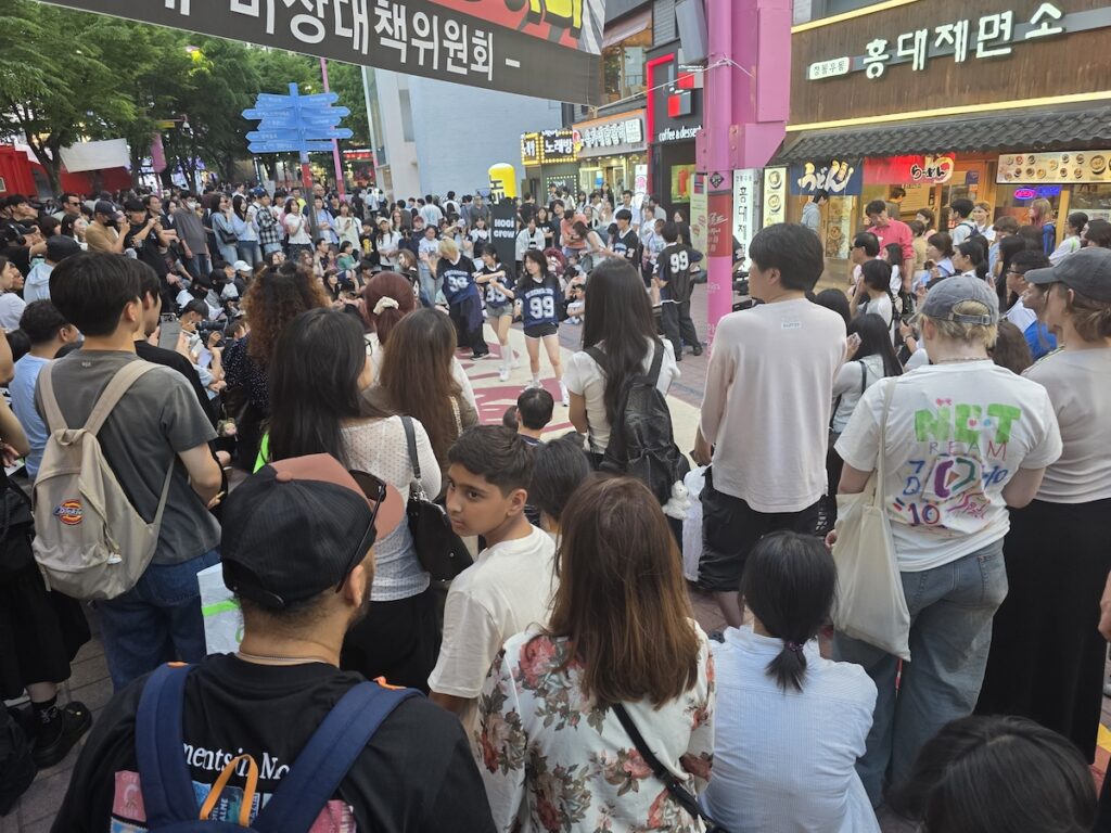 diverse-crowd-watching-dance-at-hongdae-r2-busking-zone A dynamic shot taken from within a dense crowd of diverse travelers and locals. They are all gathered tightly on a street designated as the Hongdae R2 Busking Zone to watch a K-pop cover group perform in the center.