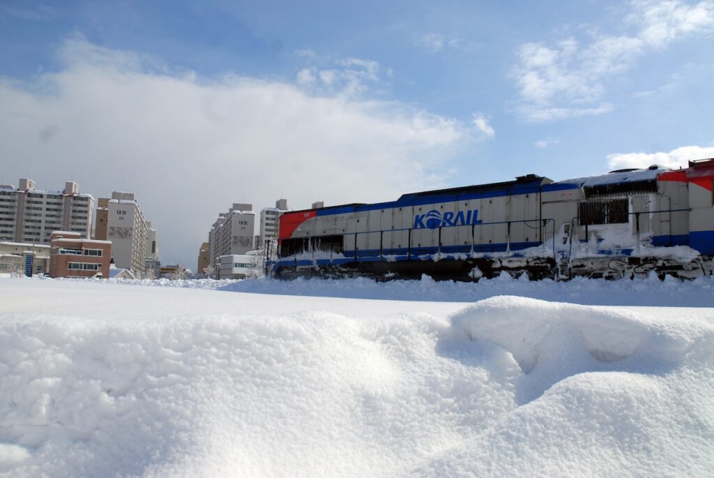 The main featured image showing a train powerfully running on a railway track completely covered in deep white snow in Donghae City, South Korea, representing the beauty of Korean winter travel.