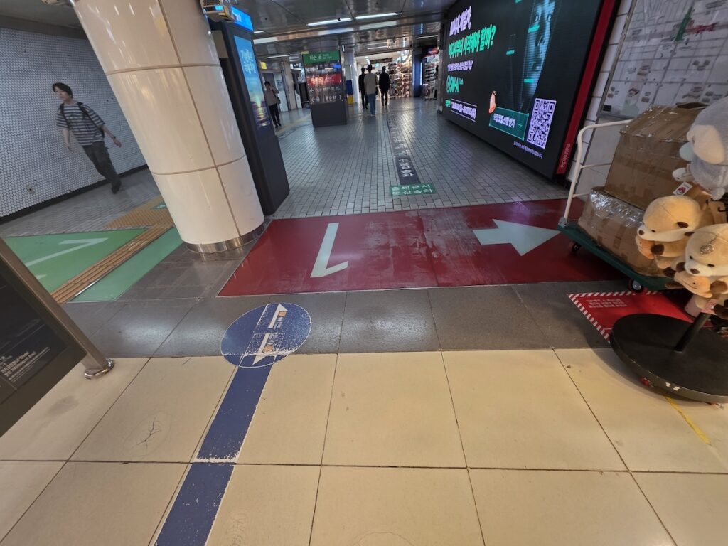 gangnam-station-underground-shopping-seoul The bustling, wide walkway of the Gangnam Station underground shopping center in Seoul with color-coded transit lines painted on the floor.