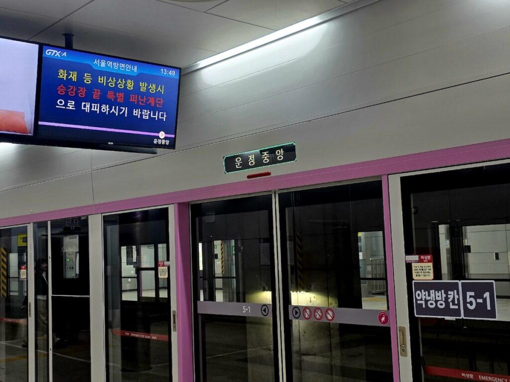 gtx-a-unjeong-central-station-b8-emergency-protocol A pink-rimmed GTX-A screen door at Unjeong Central Station with an emergency evacuation digital sign, highlighting the safety ritual for the Seoul Transportation Guide 2026.