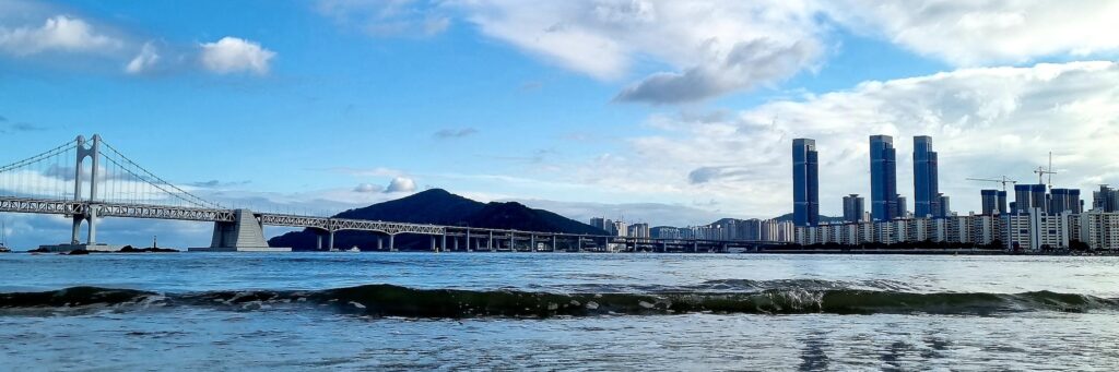 A clear morning view of Gwangalli Beach in Busan with the Gwangan Bridge under a blue sky and white clouds reflected on the wet sand.