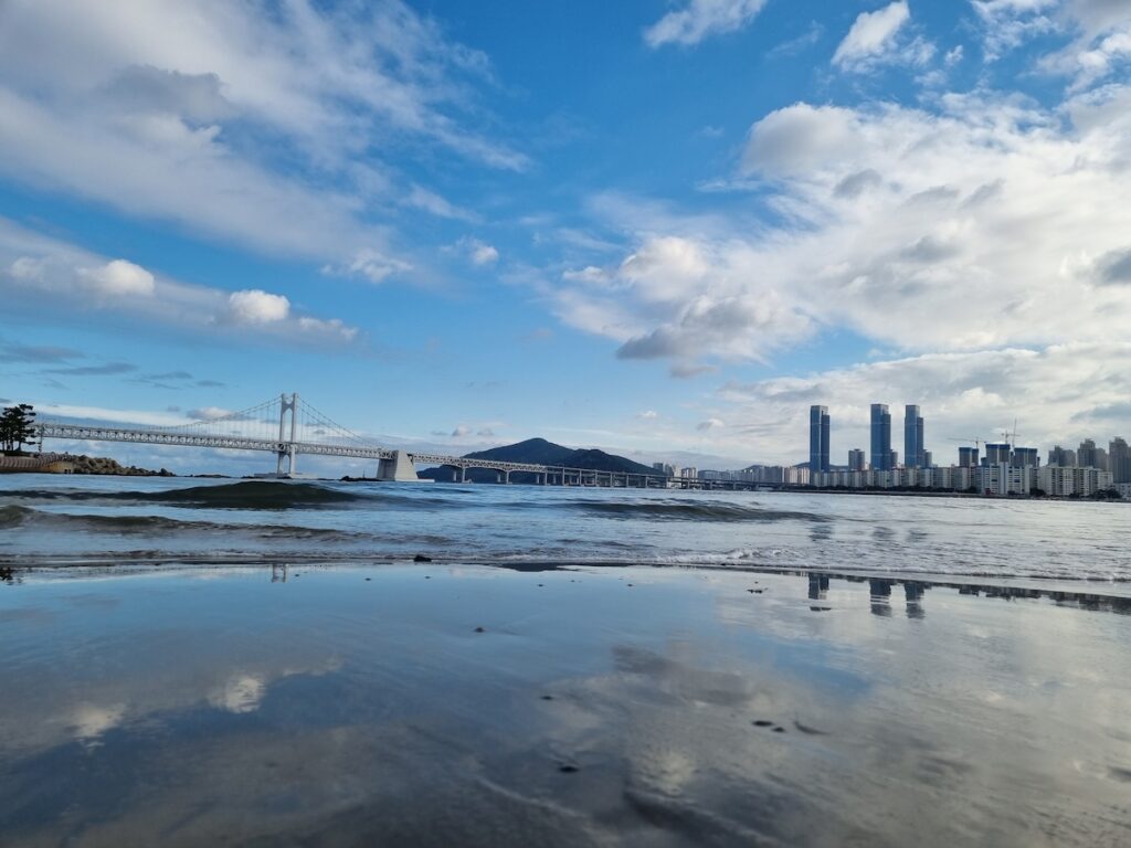 gwangalli-beach-in-busan-ocean-reflection A wide landscape view of Gwangalli Beach in Busan with gentle waves rolling in and the city skyline reflecting on the damp shoreline.