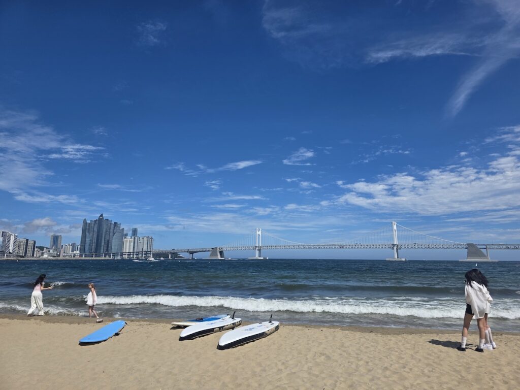 gwangalli-sup-surf-rental-tents-busan SUP and surf rental tents on Gwangalli Beach with multilingual signs and Gwangan Bridge in the distance