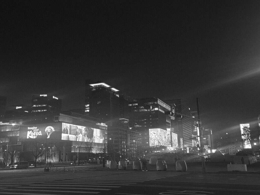 gwanghwamun-square-black-and-white-digital-billboards A cinematic black and white photograph of Gwanghwamun Square’s modern skyline, featuring bright digital billboards and "Imagine your Korea" signage, capturing the atmospheric tension of a city preparing its 5G infrastructure for a 260,000-person digital surge.