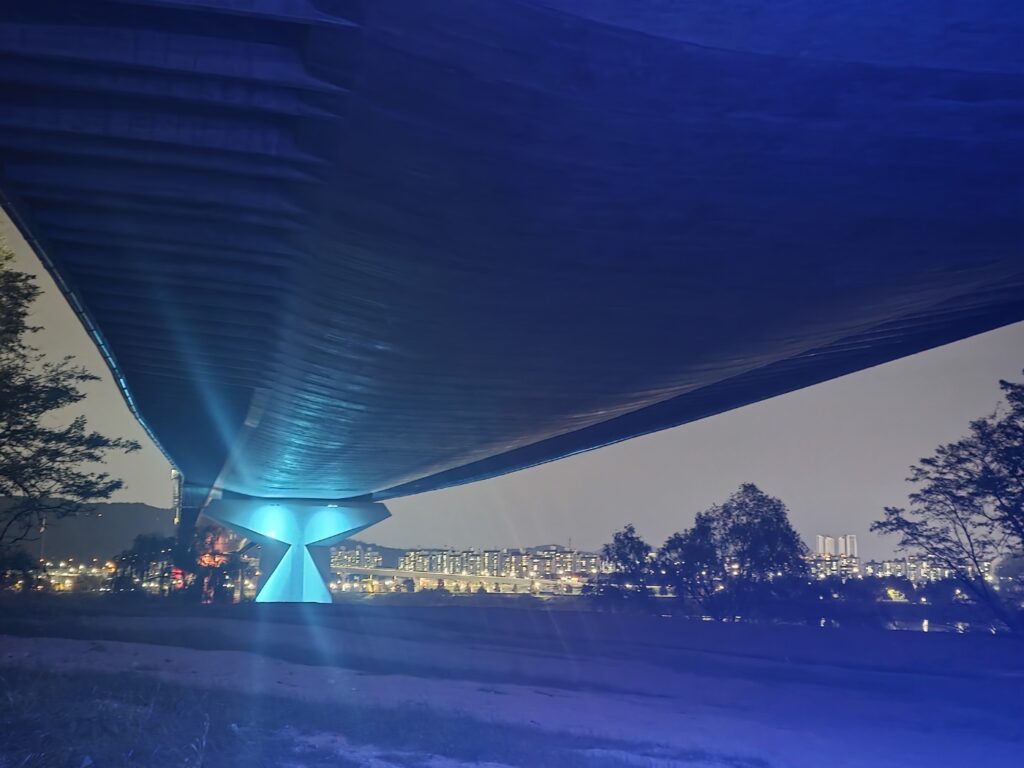 Under the Han River bridge at night in Seoul, warm lights illuminate the concrete pillars and reflect softly on the water, creating a calm and cinematic atmosphere.