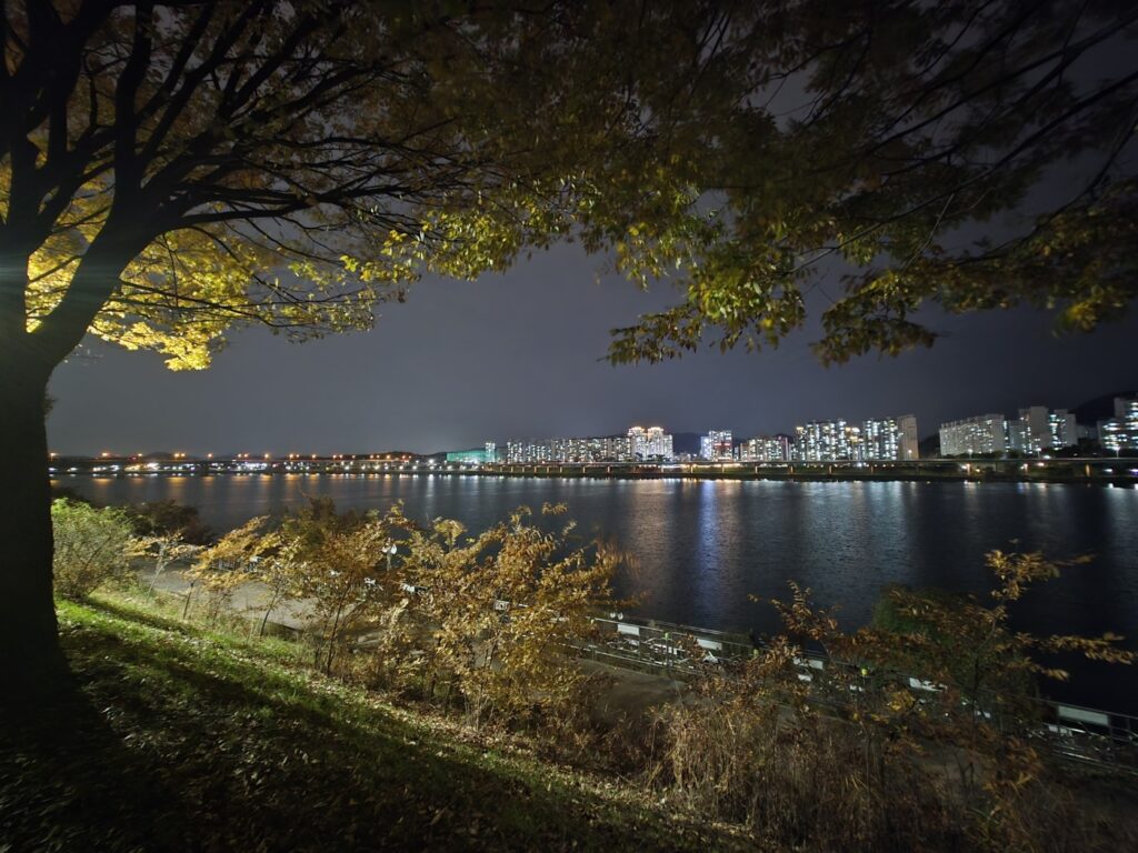 A quiet Han River walkway at night with the glowing Seoul city skyline visible across the water, symbolizing the contrast between calm and urban energy.