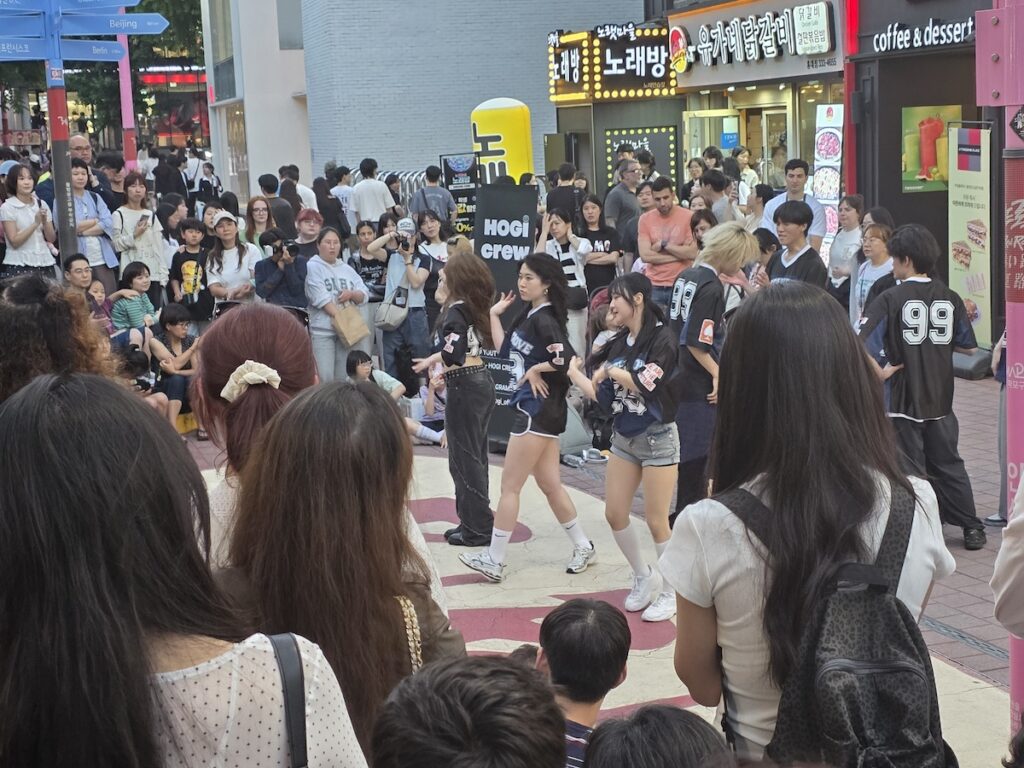 hogicrew-kpop-performance-hongdae-r2-busking-zone A close-up of a K-pop dance group, HOGI CREW, in uniform shirts, performing on the painted street of the Hongdae R2 Busking Zone, surrounded by a close circle of spectators holding up phones.