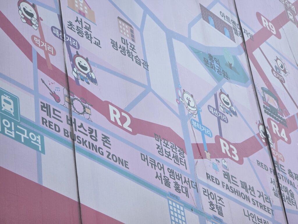 hongdae-r2-busking-zone-construction-protest-map A close-up view of a weather-beaten urban guide map in Seoul. The map features pink lines and explicitly marks 'R2 RED BUSKING ZONE' and 'R1'. Below the map, large protest banners with black and white Korean text cover the wall.