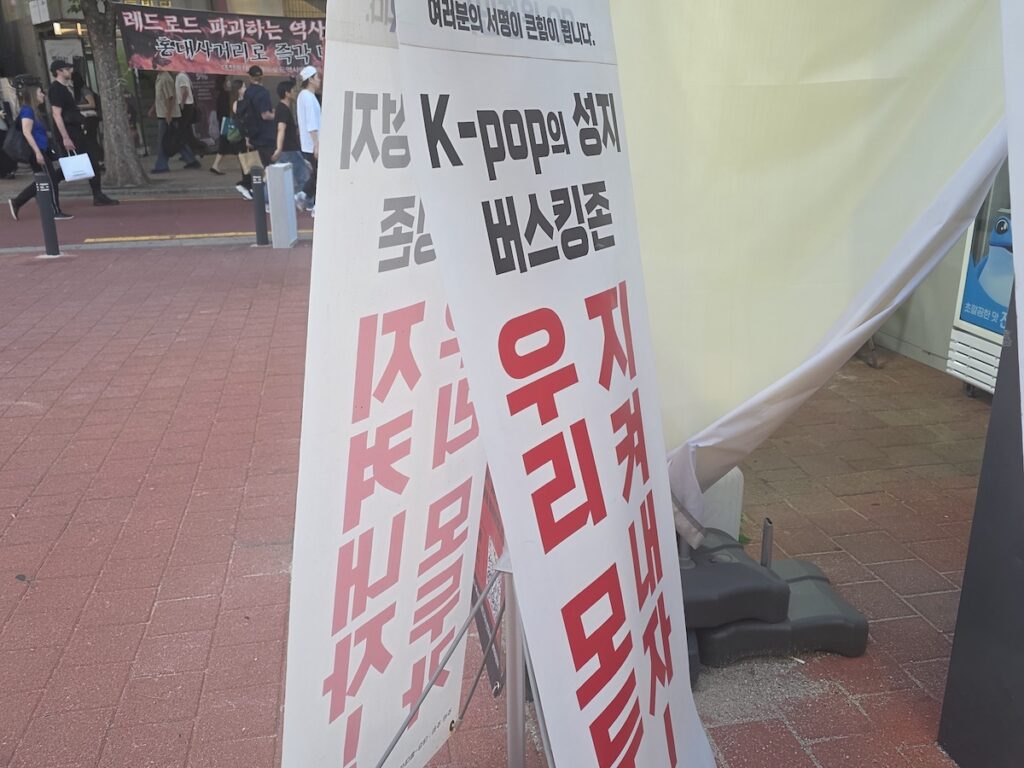 hongdae-r2-busking-zone-protest-sign A close-up of a white A-frame protest sign placed in the Hongdae R2 Busking Zone. The sign features bold black and red Korean text advocating for the preservation of the K-pop busking zone, with a blurred background showing passersby and other protest banners along the Red Road.