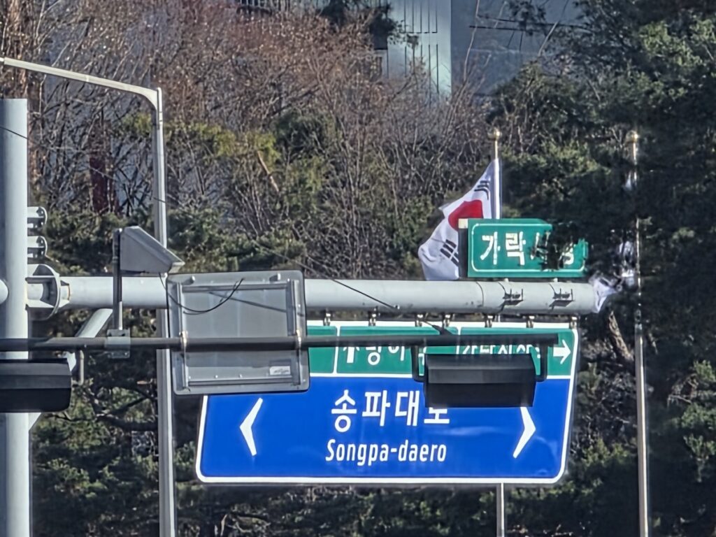 A blue Korean road sign at an intersection showing Songpa-daero, used as a primary way to report to 112 and 119 for a precise emergency location in Korea.