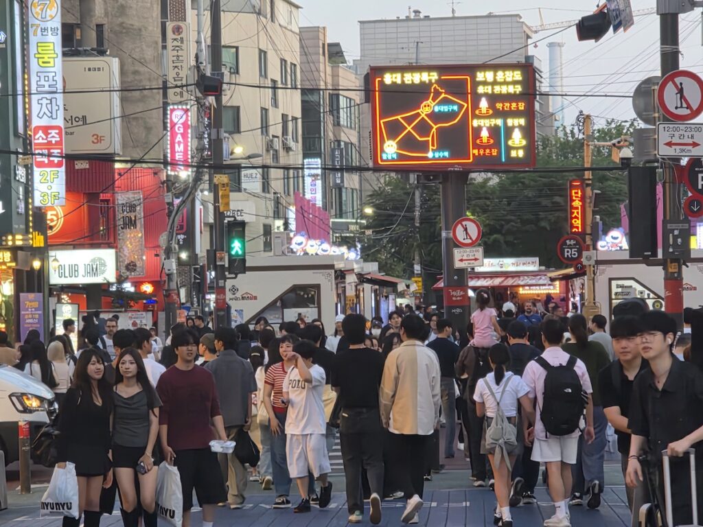 is-hongdae-too-crowded-evening-street A crowded evening street in Hongdae with shops, signs, and visitors, showing why people wonder is Hongdae too crowded