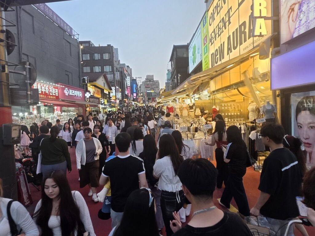 is-hongdae-too-crowded-shopping-street A busy Hongdae shopping street filled with people, shops, and bright signs, showing is Hongdae too crowded for first-time visitors