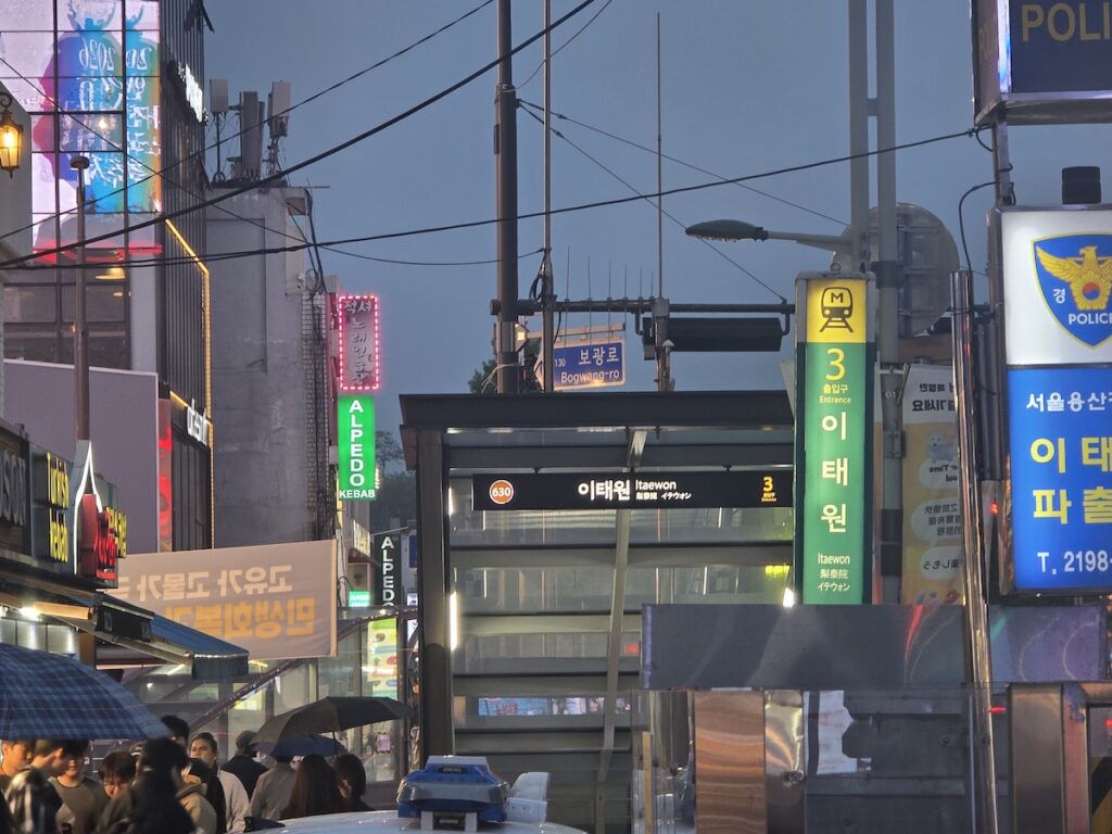 Itaewon Seoul station entrance at night with neon signs, Bogwang-ro direction, police box, and evening crowds