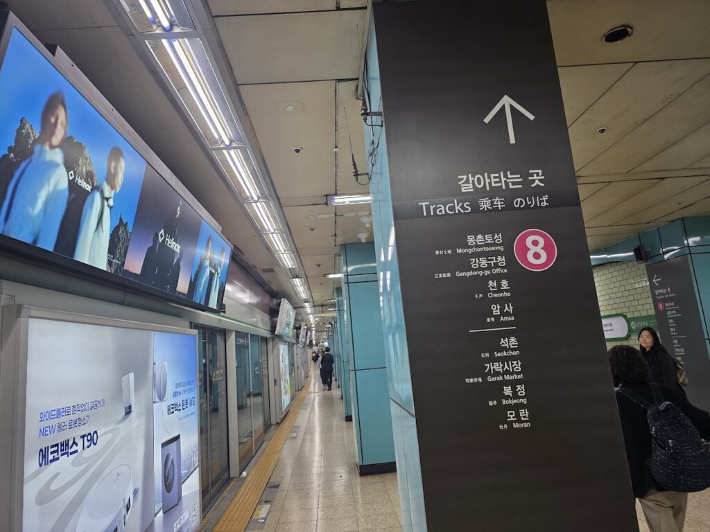 jamsil-station-line-2-platform-transfer-to-line-8-sign A wide-angle view of a Seoul Subway Line 2 platform at Jamsil Station. The platform is brightly lit with LED linear lights and lined with glass screen doors featuring large commercial advertisements. In the foreground, a prominent dark grey rectangular pillar features a large white upward-pointing arrow and the text 'κ°μνλ κ³³' (Tracks) in multiple languages. Below this, a comprehensive list of Line 8 stations is provided in vertical order, starting from Mongchontoseong and ending at Moran, with the pink Line 8 circle symbol adjacent to the station names. Commuters are walking down the clean tiled platform toward the transfer path indicated by the arrow.