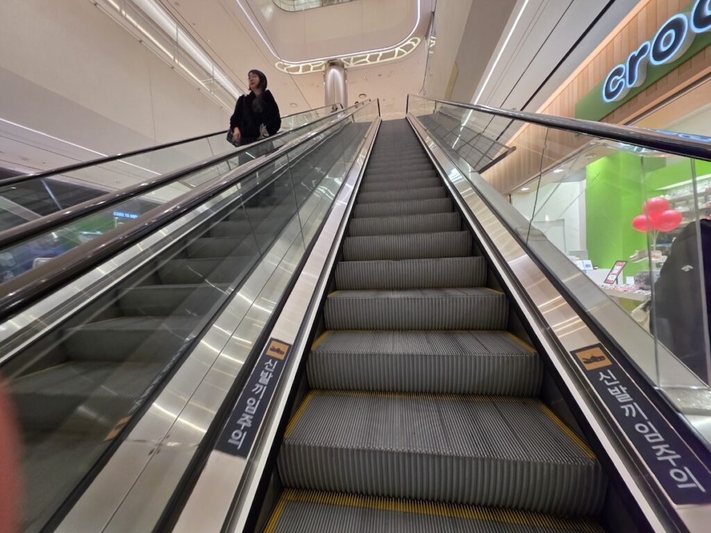 jamsil-station-transfer-escalator-safety-sign Escalator inside Lotte World Mall during a Jamsil Station Transfer, highlighting the Korean safety warning 'Caution: Watch your shoes'.