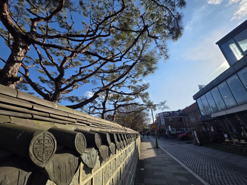 jeonju-hanok-village-traditional-stone-wall-pine-tree A cinematic view of a traditional Korean stone wall (Damjang) and pine trees under a clear blue sky in Jeonju Hanok Village, representing the serene lifestyle of the city.