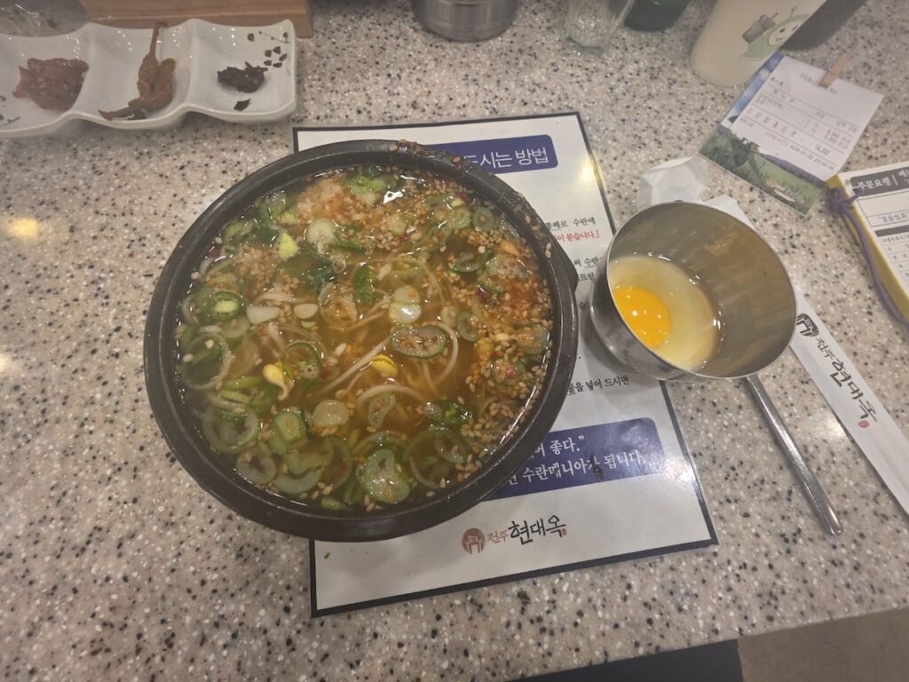 jeonju-kongnamul-gukbap-hyundaeyok-breakfast-guide A top-down view of a steaming bowl of Jeonju Kongnamul Gukbap (bean sprout soup) served with a side of poached egg (Suran) at a local restaurant, an essential experience in the **Jeonju Travel Guide**.