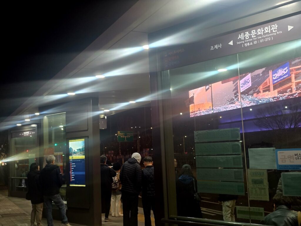 korea-points-based-visa-system-bus-stop-waiting Professionals waiting at a modern Seoul bus stop featuring traditional art, representing the proactive waiting period within the D-7 to F-2-7 visa Korea roadmap.