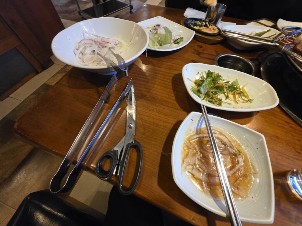 korean-bbq-tools-scissors-tongs-onions Traditional Korean BBQ dining tools including stainless steel scissors and tongs next to sliced onions in soy-based dipping sauce