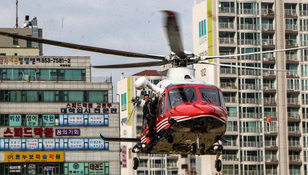 korean-firefighting-helicopter-2025 "A Korean firefighting helicopter hovering over a forest area, releasing water to extinguish a wildfire during an emergency operation in South Korea."