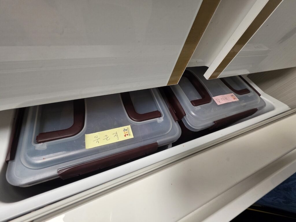 korean-kimchi-refrigerator-containers-mugeunji Inside a Korean kimchi refrigerator drawer showing airtight containers labeled with Mugeun-ji (aged kimchi) and Altari kimchi.