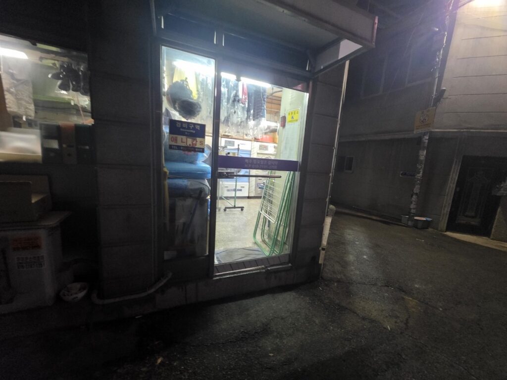 A brightly lit neighborhood laundry shop in a quiet Seoul alley at night, showing the warm and welcoming entrance of a traditional Korean setakso.