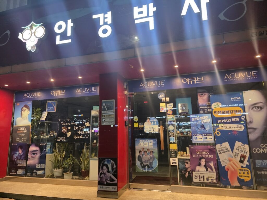 A brightly lit Korean optical shop at night showing its neon signs, emphasizing the convenience of late-night glasses shopping.