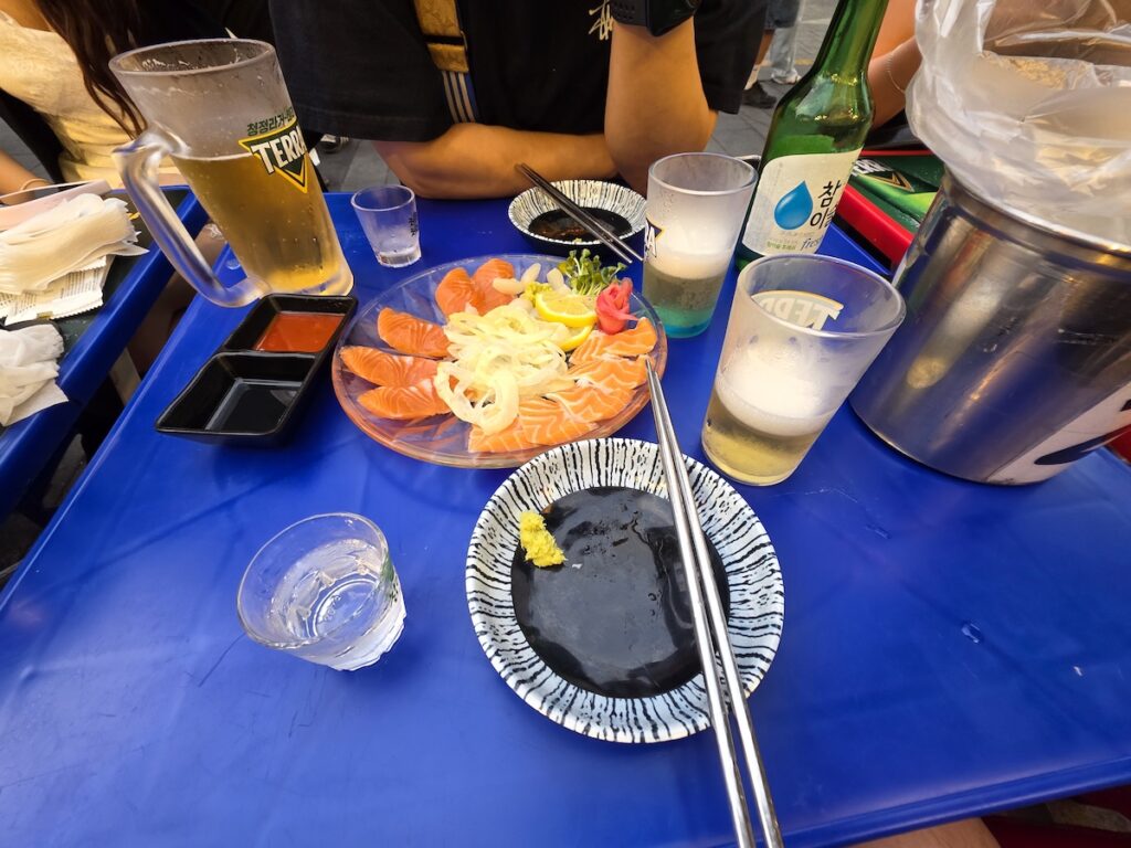 korean-outdoor-drinking-culture-salmon-soju-beer Salmon sashimi with soju and beer on a blue street table, typical of Korean outdoor drinking culture