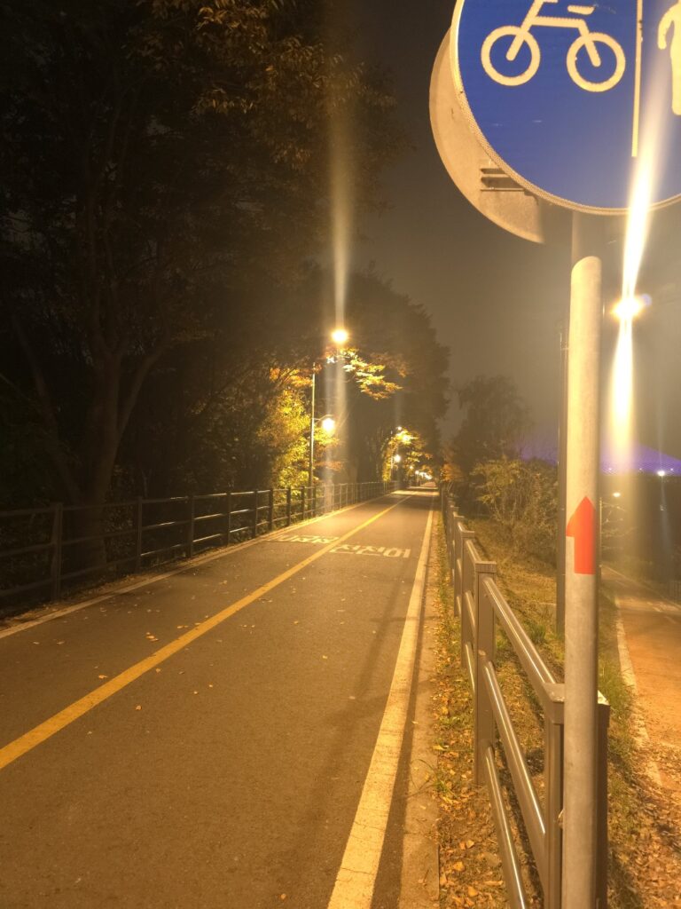 A quiet, empty walkway along the Han River at night in Seoul, illuminated by bright streetlights that create a feeling of calm solitude and safety.