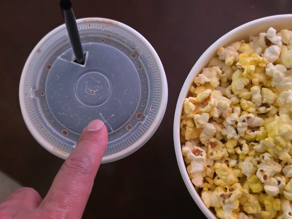 lotte-cinema-jamsil-dual-straw-cup-lid-ritual A detailed top-down view of a Lotte Cinema Jamsil soda cup lid with dual straw apertures designed for the unique coupled-centric Korean Multiplex Culture next to a bucket of popcorn.