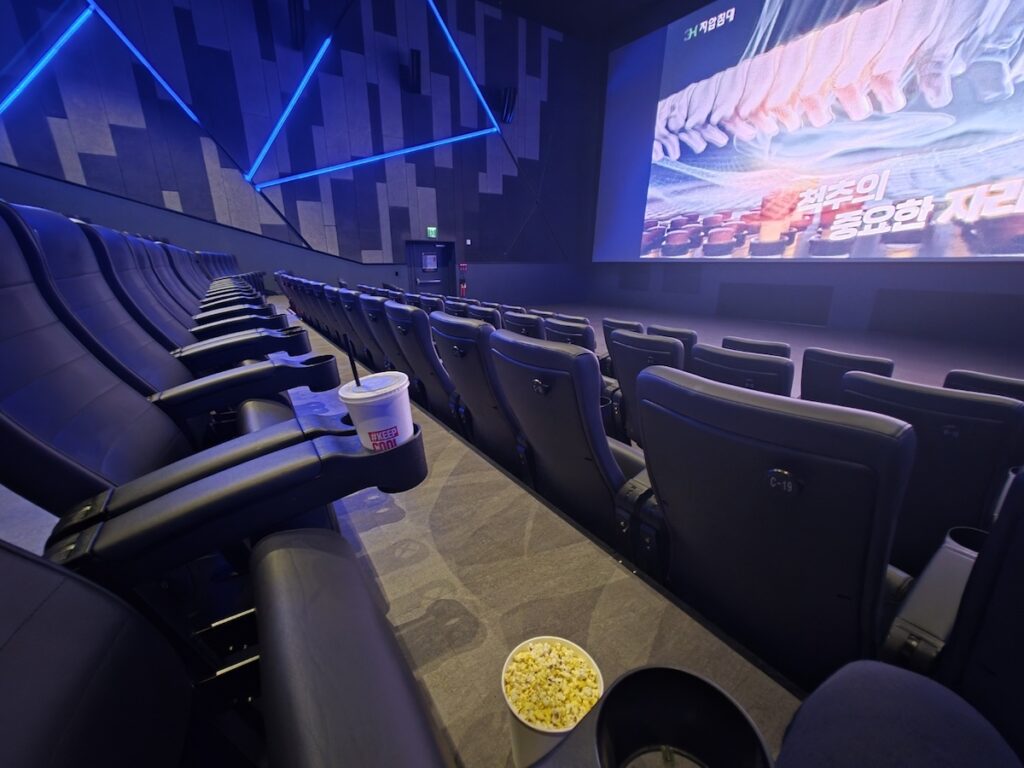The interior of a Lotte Cinema Jamsil screening room with reclinable seating and a large screen displaying advertisements before the main event as part of the Cinematic Seoul experience.