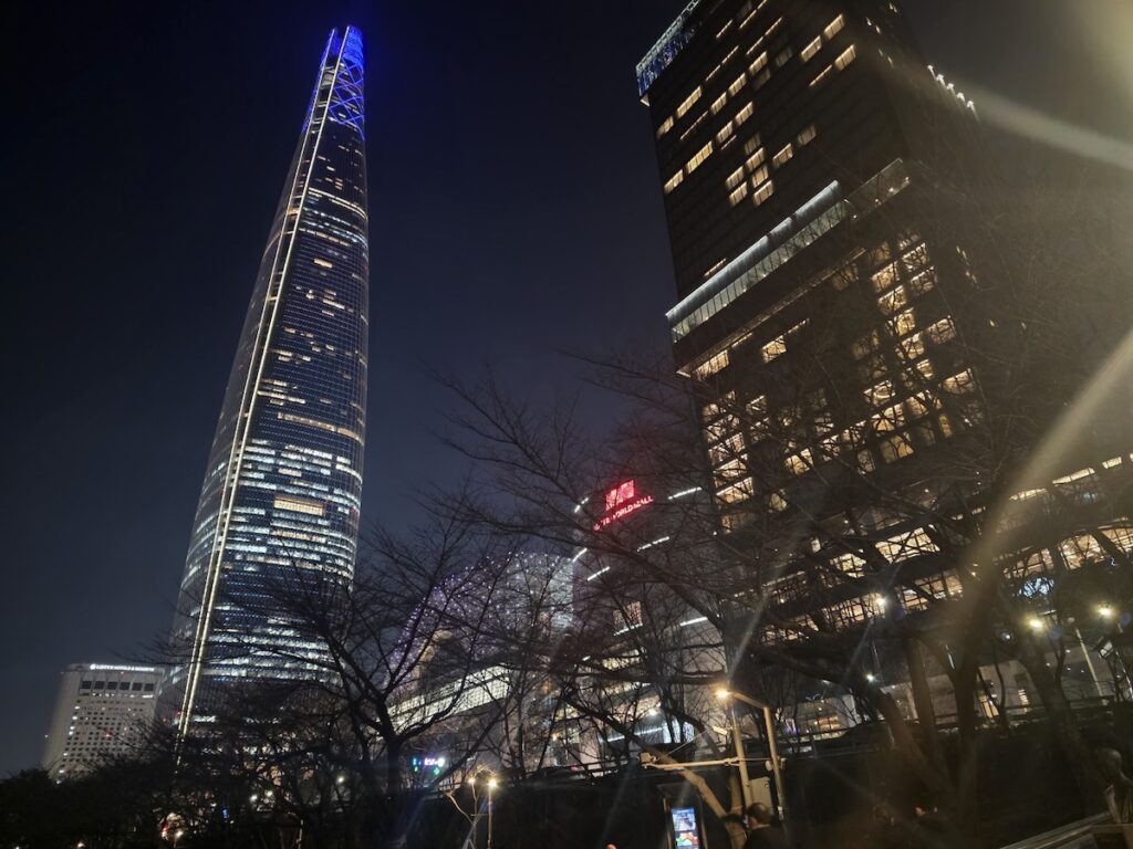 A cinematic low-angle shot of the glowing Lotte World Tower and surrounding skyscrapers at night for the **Best Seoul Night View Spots 2026**.