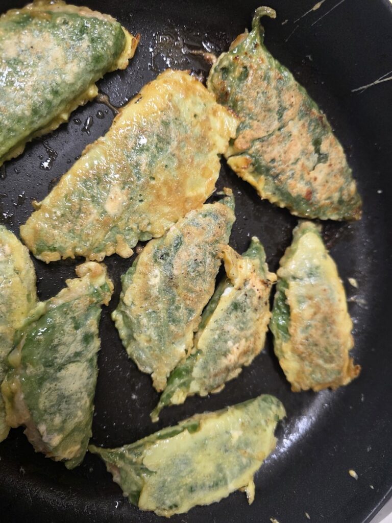 A Korean home kitchen scene showing hands pan-frying assorted jeon (Korean pancakes) on a skillet during Chuseok holiday preparations.