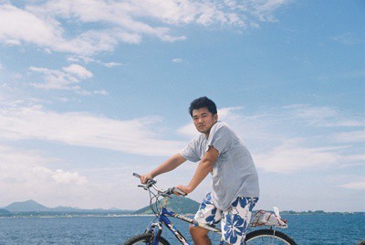 marcus-cycling-jeju-island-coastal-road. Marcus Park riding a bicycle along the blue coastal road of Jeju Island under a clear sky, representing a personal travel experience in Korea.