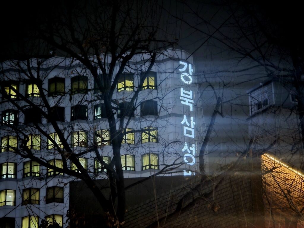 The neon sign of Kangbuk Samsung Hospital seen through bare tree branches at night, symbolizing the distant and cold nature of healthcare during a Medical Emergency Korea Without ARC.