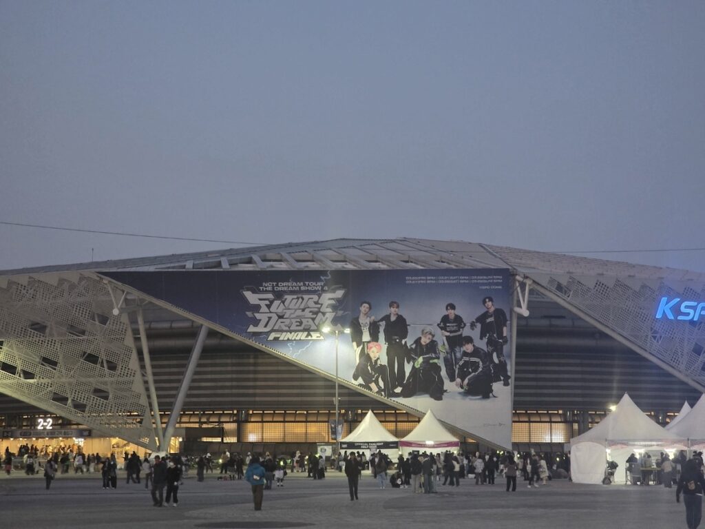 nct-dream-seoul-concert-2026-gate-2-1 A nighttime shot of "GATE 2-1" at the KSPO DOME during the NCT DREAM Seoul Concert 2026. The image shows a long, organized queue of fans waiting under the bright white lights of the entrance, with a large blue "GATE 2-1" sign clearly visible above the crowd.
