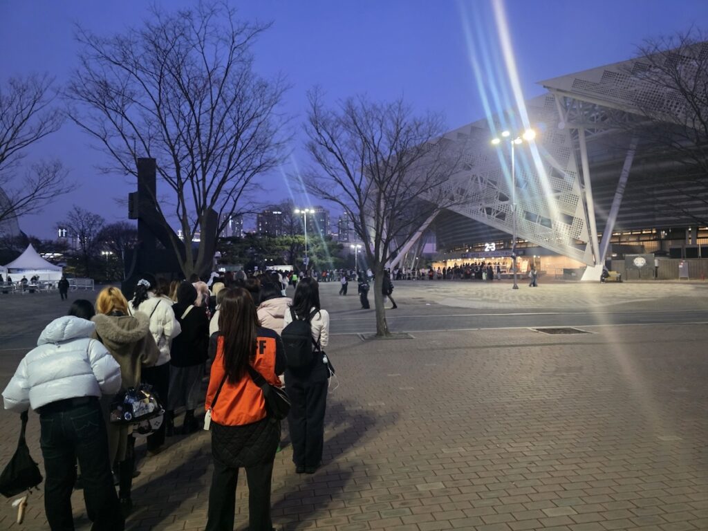 nct-dream-seoul-concert-2026-stadium-exterior A shot of the arena's curved glass exterior at night. A few fans are seen resting or waiting on benches in the foreground, with the massive, glowing stadium in the background, capturing the quiet moments before the NCT DREAM Seoul Concert 2026 performance starts.